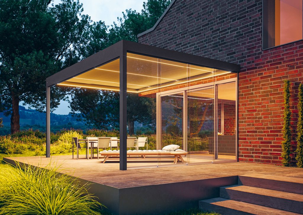 Modernes Terrassendach mit Glas-Schiebeelementen und integrierter LED-Beleuchtung, hochwertige Pergola an Einfamilienhaus für wettergeschützten Outdoor-Wohnraum und stilvolle Terrasse