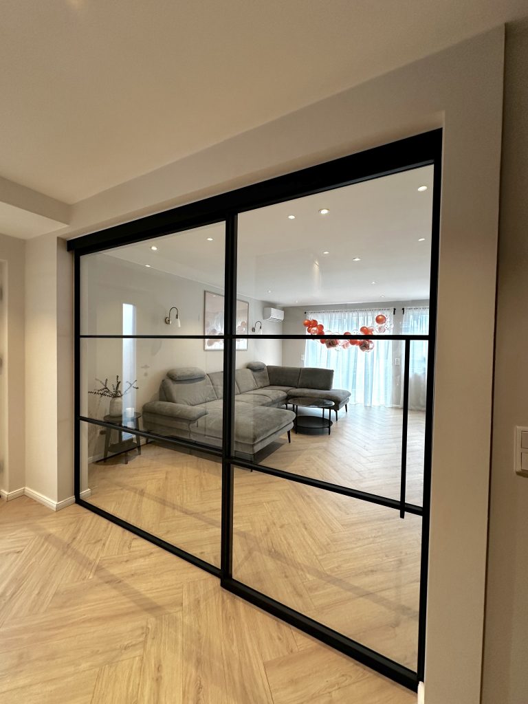 alt="Zweiflügelige Lofttür mit schwarzem Stahlrahmen und Klarglas als moderner Raumteiler im Wohnbereich – maßgefertigte Lofttüren für Trier, Konz und Luxemburg"